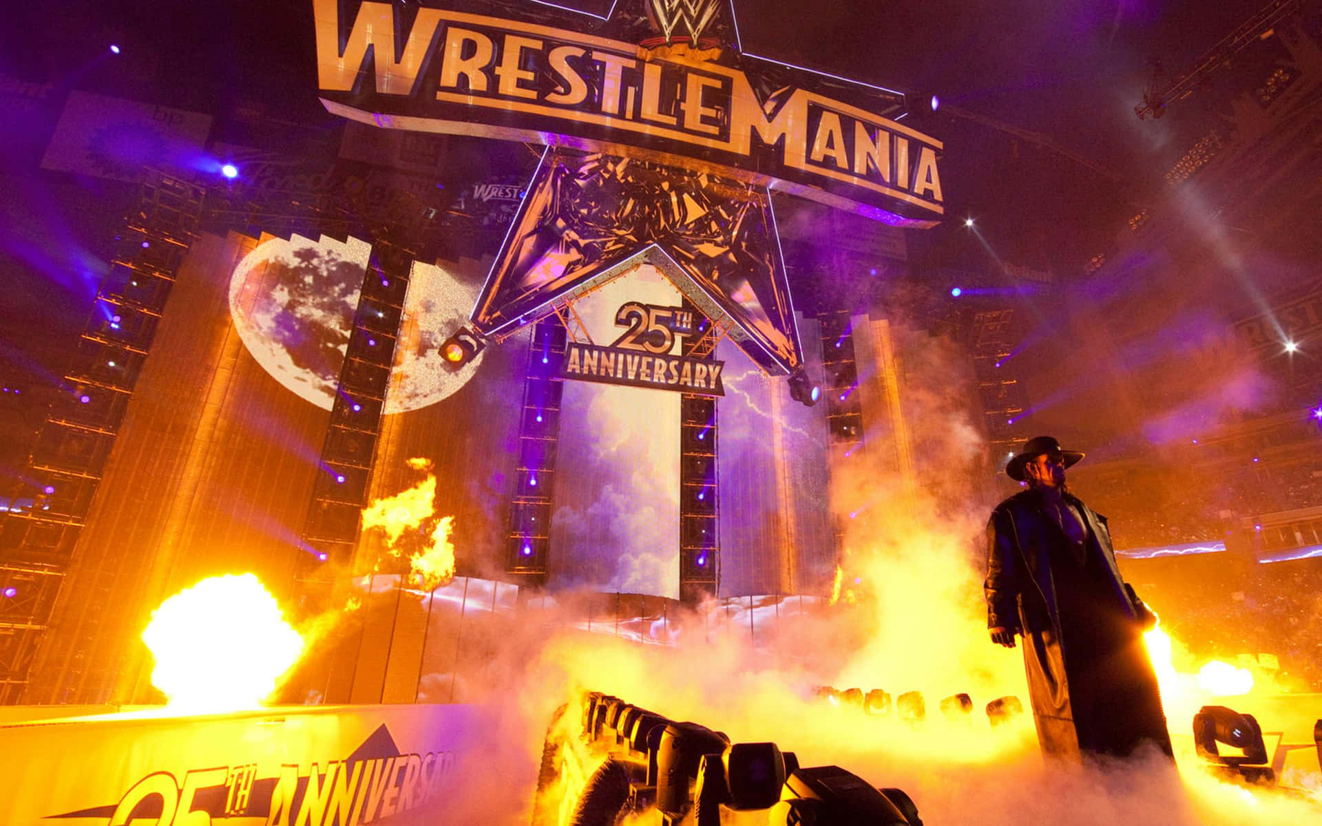  壁紙 — 一位穿著長外套的摔角手站在WrestleMania 25週年紀念舞台下，周圍是熾熱的爆炸，伴隨著戲劇性的紫色和橙色燈光。