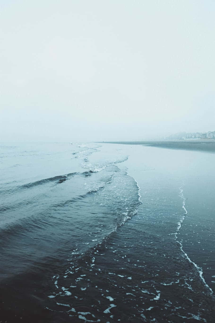 Minimalist Beach 桌布 壁紙 — 霧氣瀰漫的海浪輕輕拍打黑暗的海岸，蒼白的陰天中遠處建築物幾乎看不見。