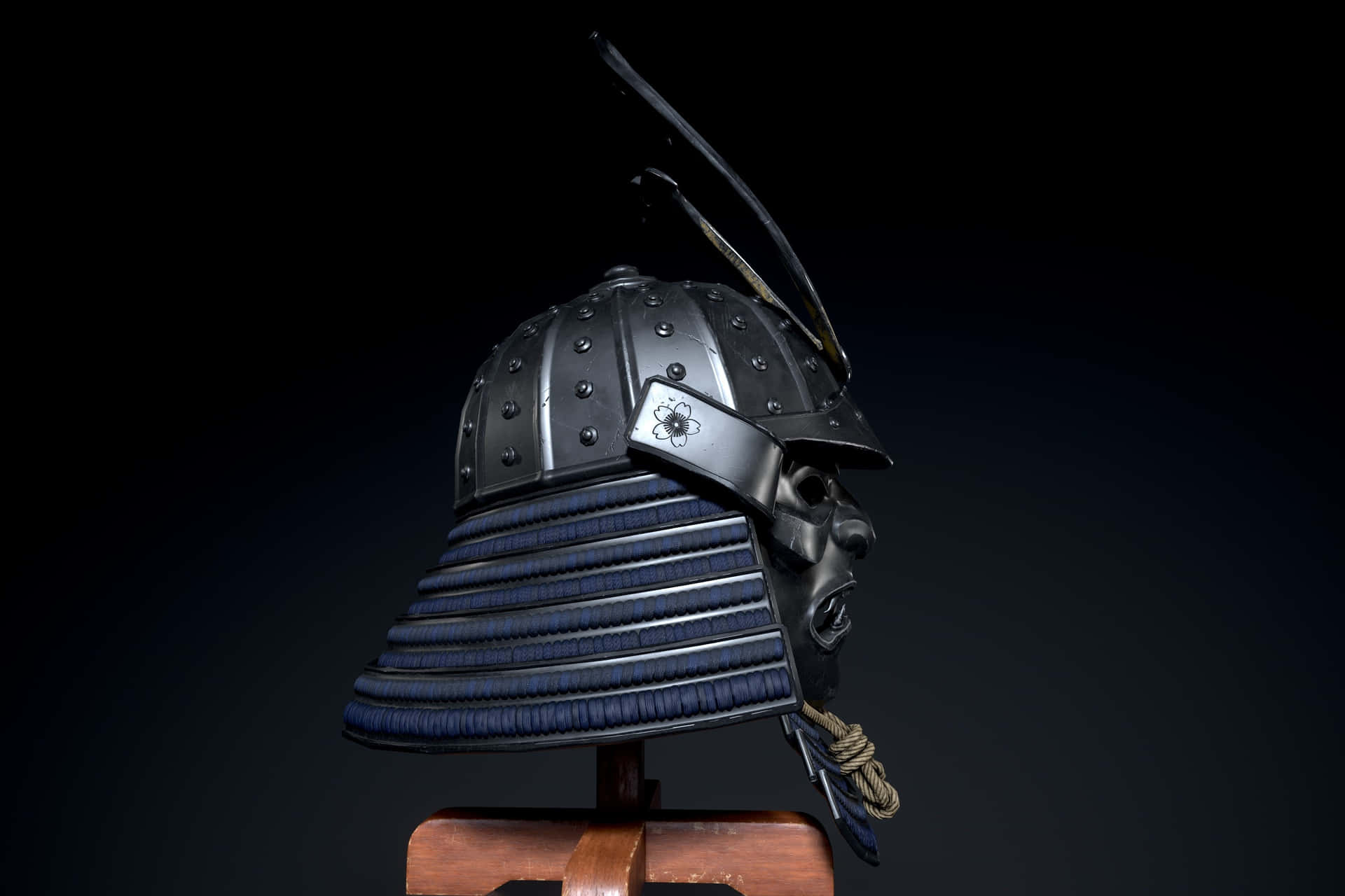  壁紙 — 一個細節豐富的武士頭盔，帶有深藍色漆面金屬板和金屬裝飾，放置在黑色背景前的木製支架上。