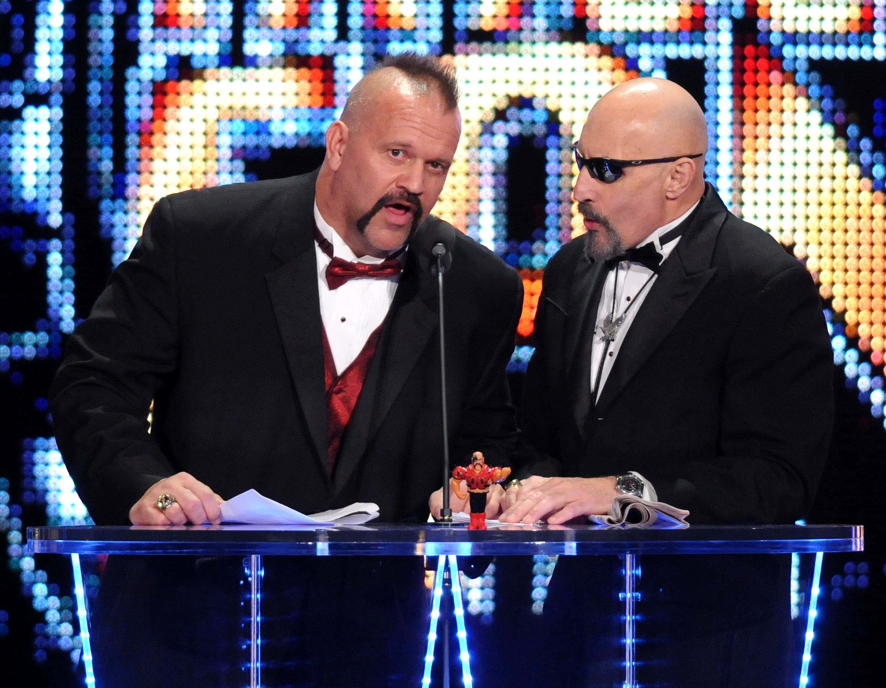WWE 2011名人堂活動中的Road Warrior Hawk 壁紙 — 兩位身穿禮服的WWE評論員靠在一張發光的藍色桌子上，背景是充滿活力的像素化背景。