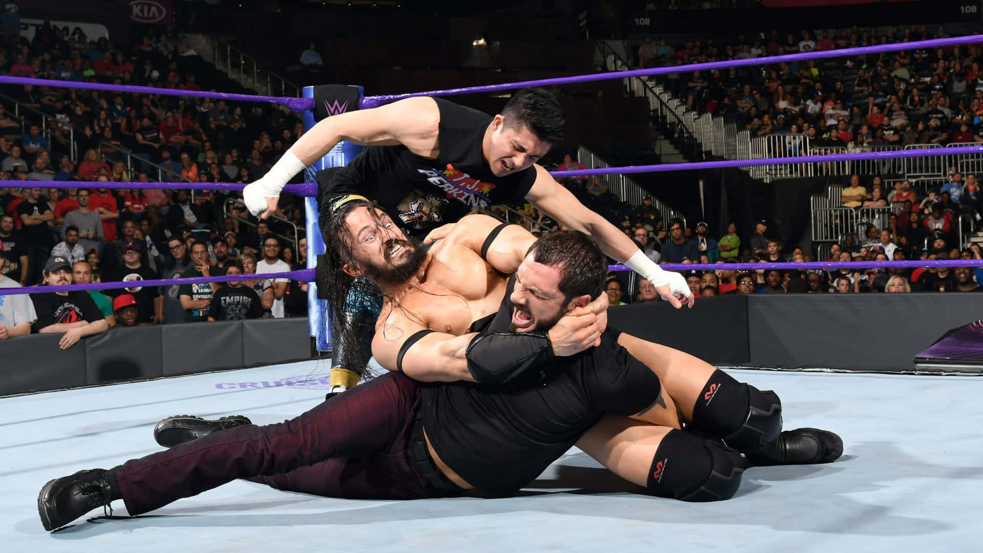 WWE 205賽事中奧斯汀·艾瑞斯遭T.J. 傑克森與內維爾夾擊 壁紙 — 三名摔角手在擂台上激烈纏鬥，全場觀眾在明亮的場館燈光下觀看。
