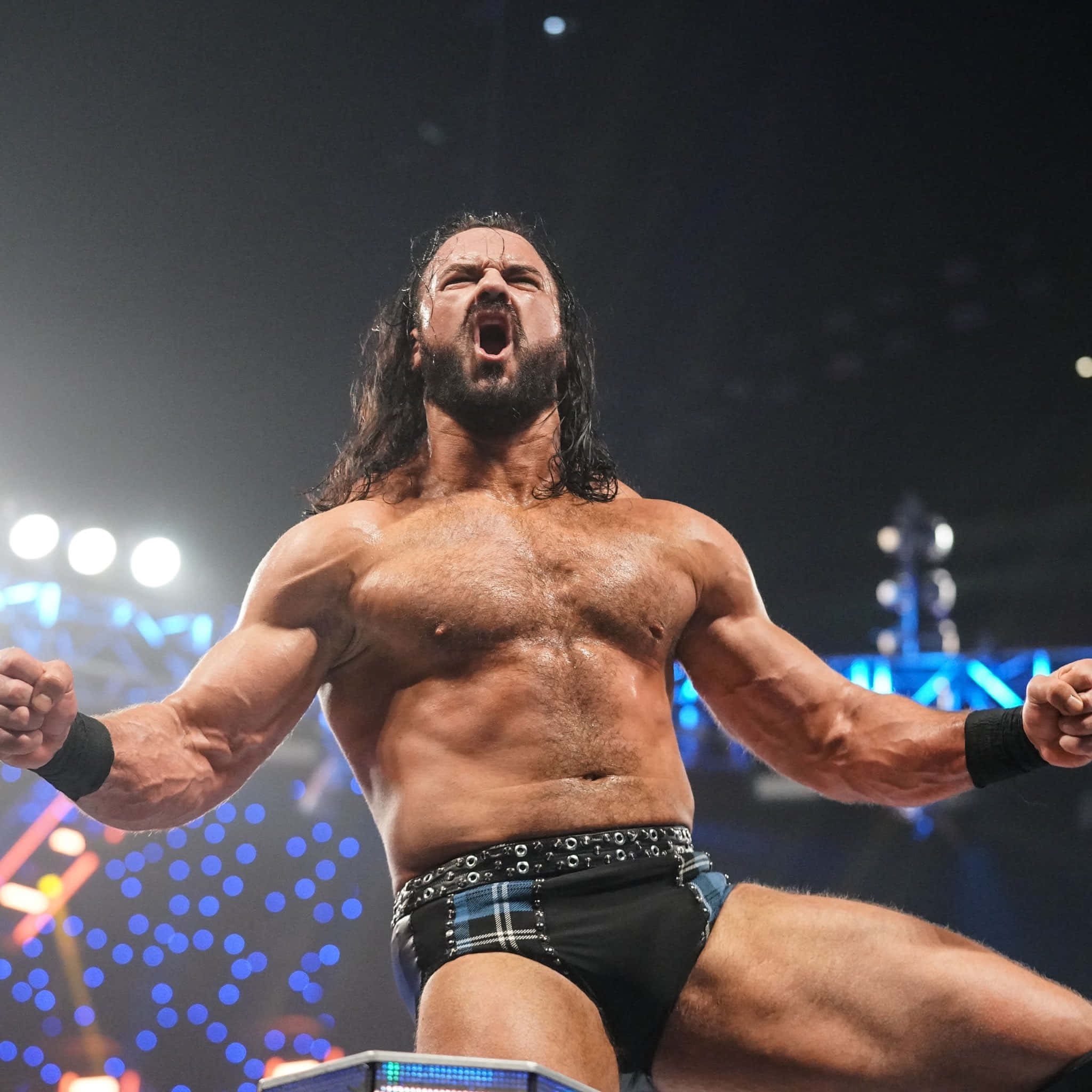 WWE專業摔角手德魯·麥金太爾震撼怒吼 壁紙 — 一位肌肉發達、留著深色長髮和鬍子的摔角手，在身後藍色燈光的明亮擂台上勝利地咆哮。