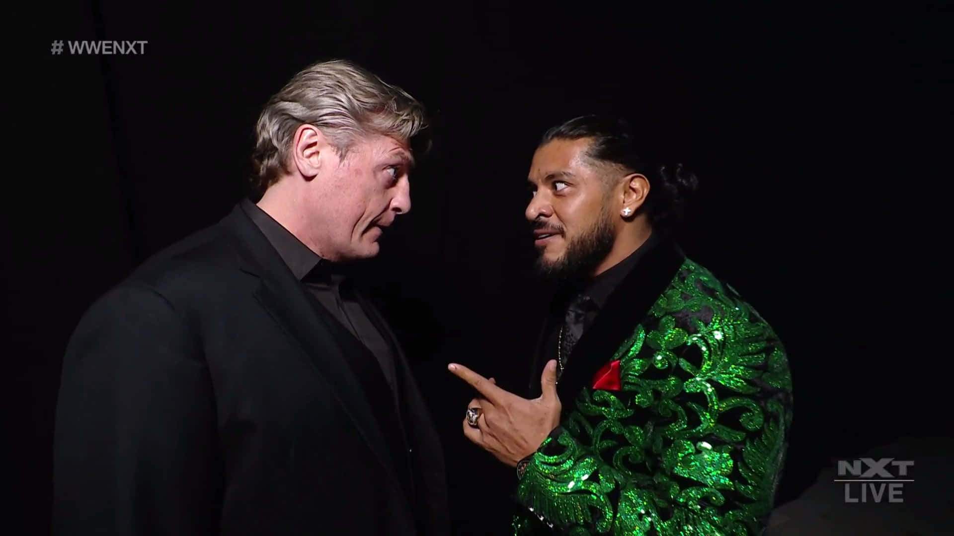 標題：WWE NXT 對決場景 - 威廉·瑞格 壁紙 — 兩位WWE摔角手在緊張時刻對峙，一人身穿黑色西裝，另一人穿著閃亮綠色外套，背景昏暗。