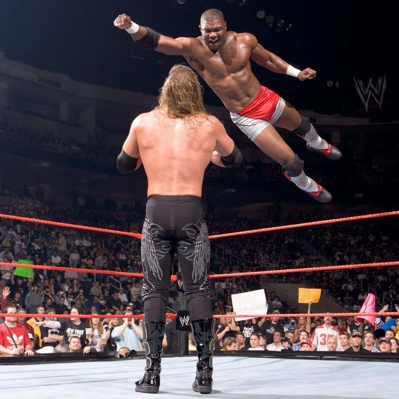 謝爾頓·本傑明在 WWE Raw 中對 Edge 施以窒息摔 壁紙 — 一位肌肉發達的摔角手在燈光明亮的擂台上，在咆哮的粉絲包圍下，凌空撲向對手。