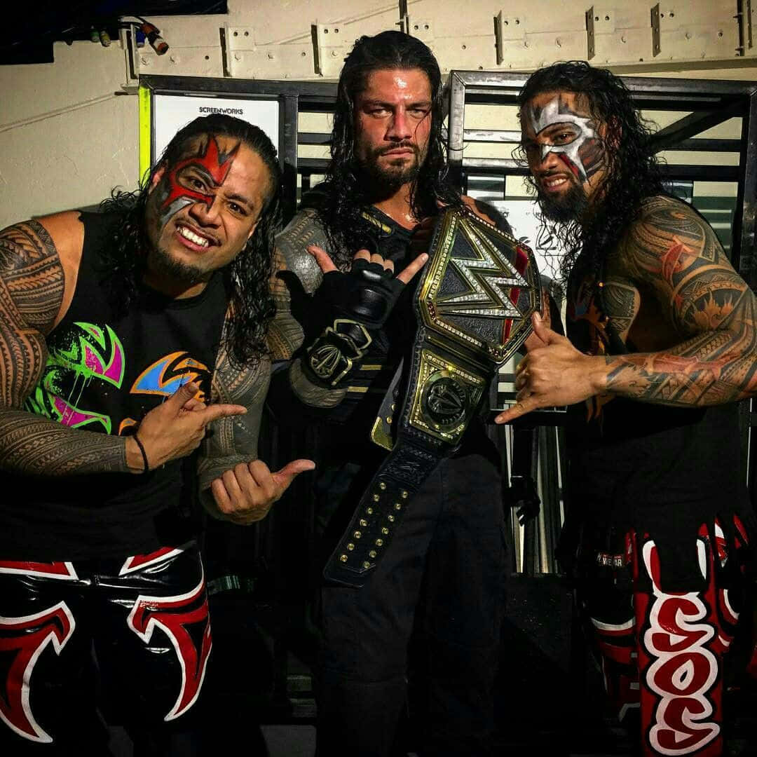 WWE Roman Reigns 與 Jimmy 與 Jey Uso 壁紙 — 三位紋身摔角手帶着臉部彩繪，在後台區域自信地擺姿勢，手持冠軍腰帶。