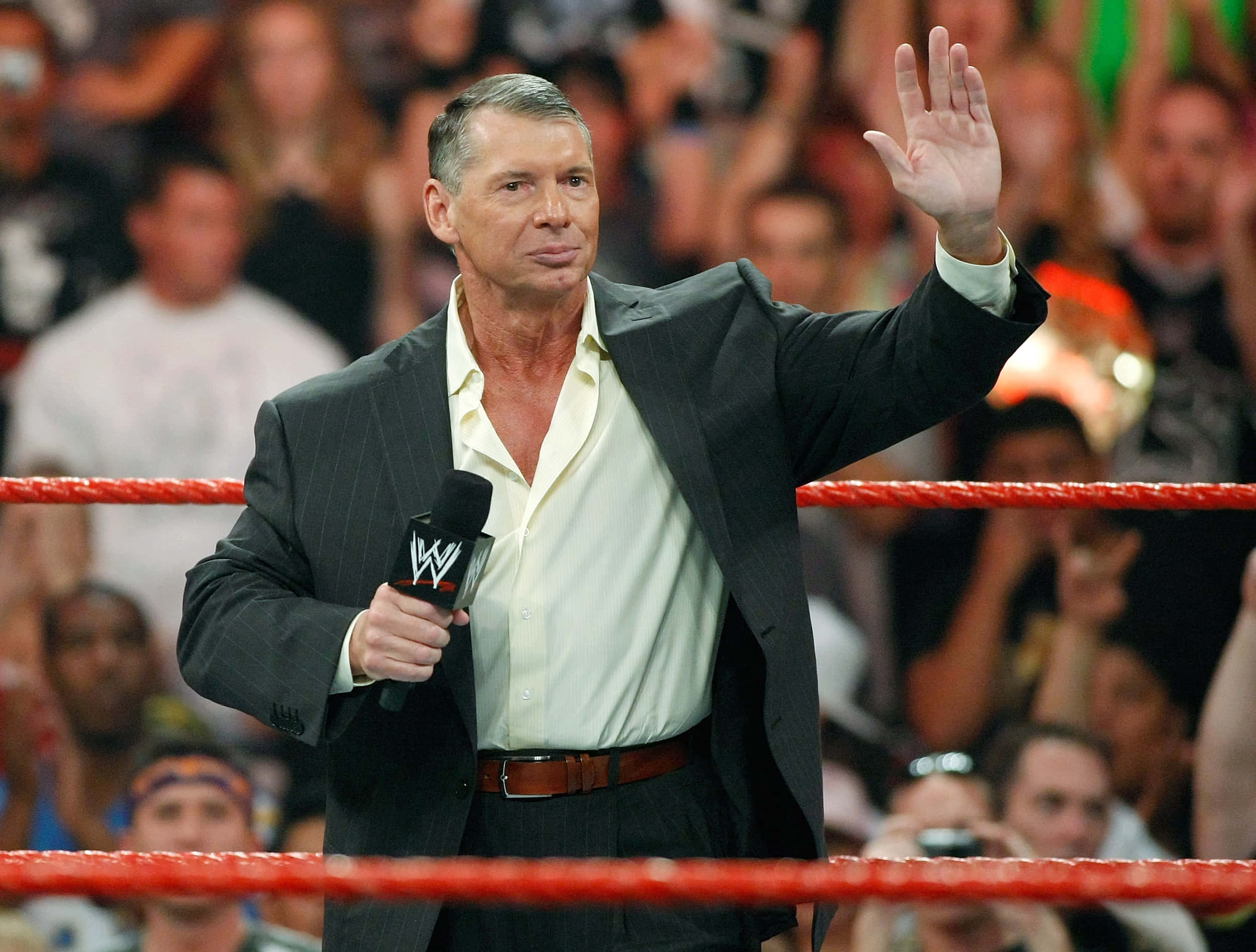 WWE主席文斯·麥馬漢高興向觀眾揮手 壁紙 — 資深摔角評論員 Vince McMahon 在燈光明亮、帶有紅色繩索的摔角場內向粉絲揮手。