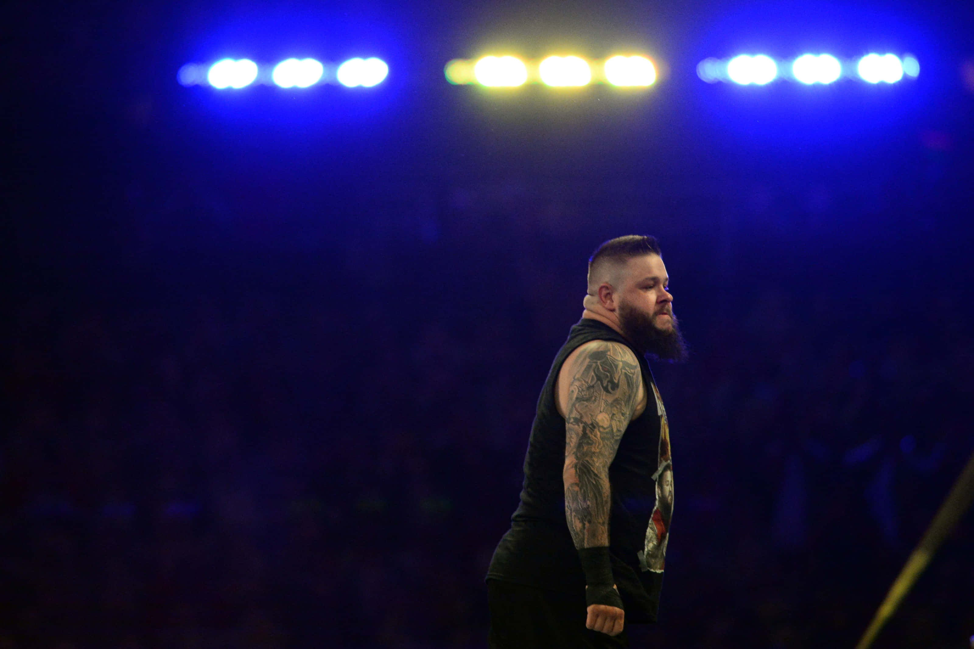 WWE 摔角手凱文·歐文斯 壁紙 — 一位留鬍鬚、有紋身的摔角手在明亮的藍色和黃色場館燈光下以側面站立。