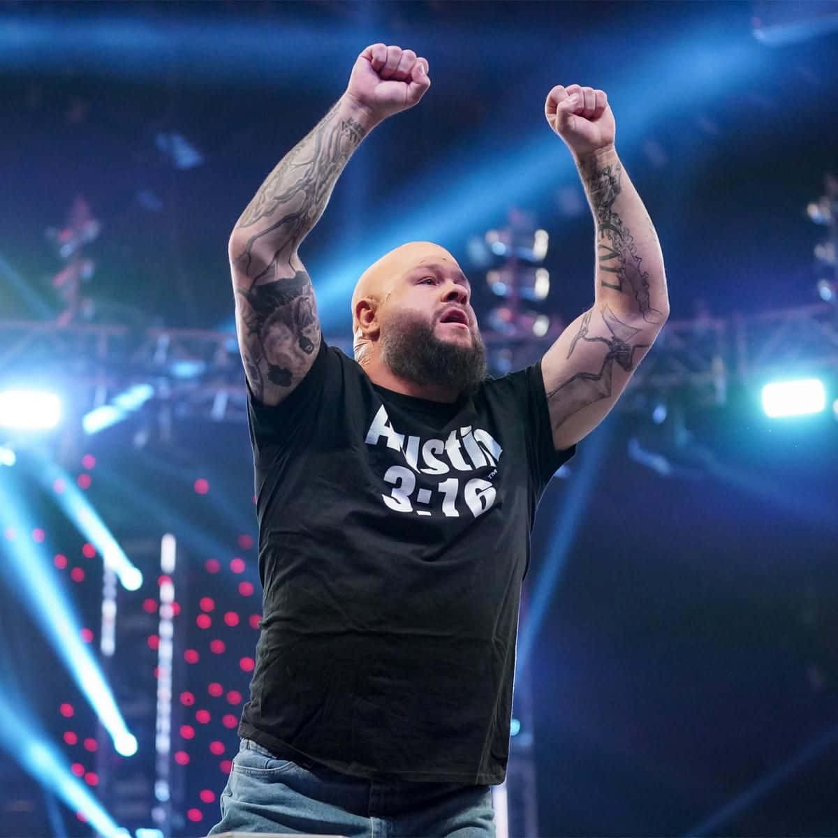 WWE 摔角手凱文·歐文斯於 RAW 賽事中 壁紙 — 一位光頭鬍鬚、手臂有紋身的摔角手在明亮的藍色舞台燈光下勝利地舉起拳頭。