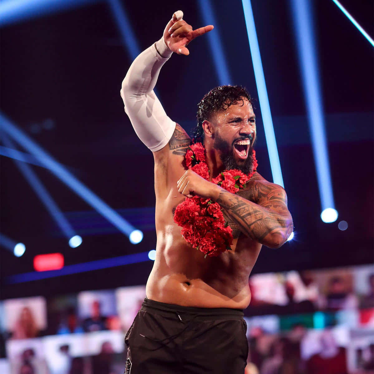 WWE 明星 Jey Uso 配戴紅色花項鍊 壁紙 — 一位肌肉發達、有紋身、戴著紅色花環的摔角手，在明亮的藍色舞台燈光下勝利地吶喊。