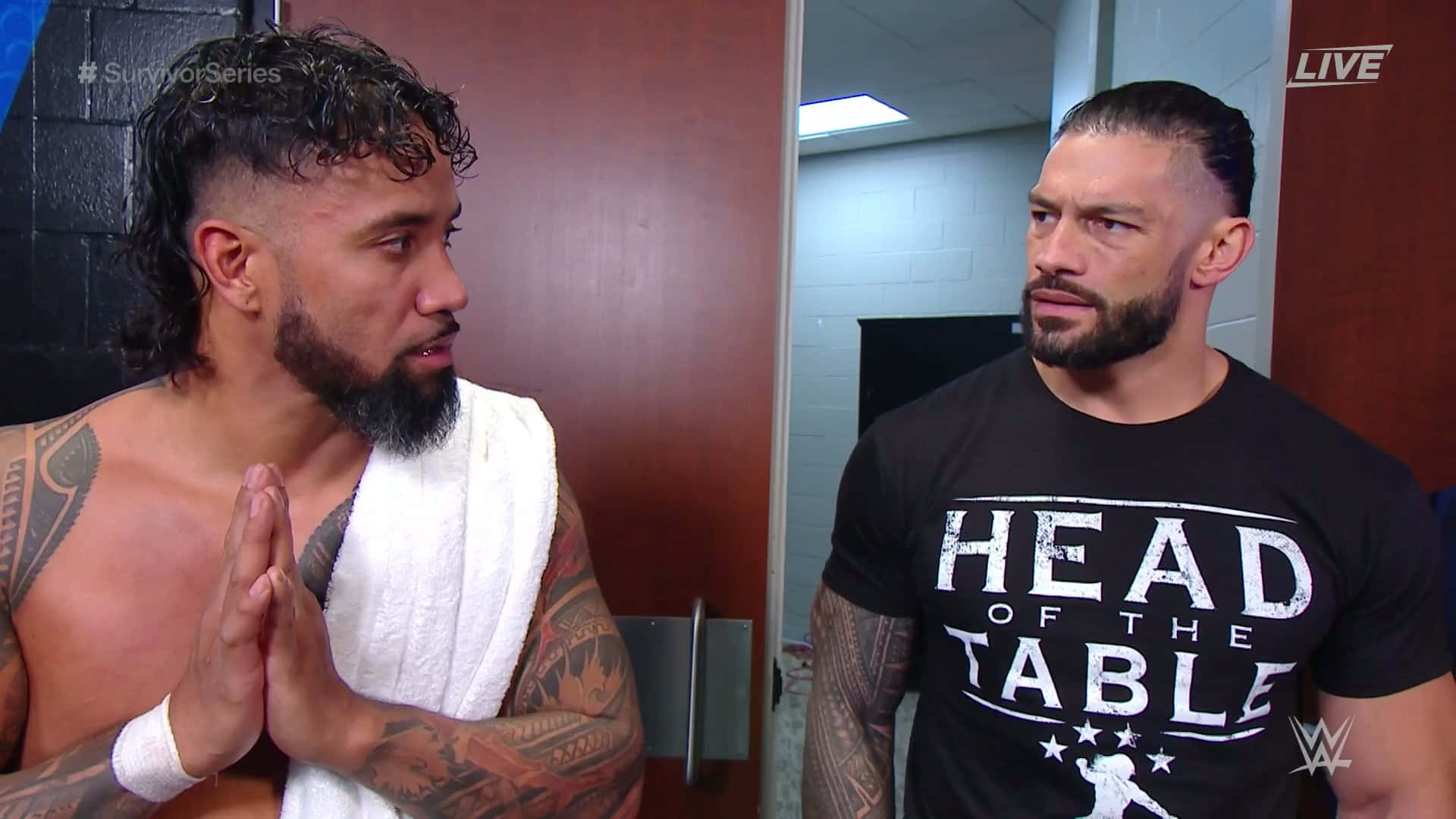 WWE 明星：傑伊·烏索與羅曼·雷恩斯現場演出 壁紙 — 兩名肌肉發達的摔角手在後台走廊對峙，一人赤裸上身披著毛巾，另一人穿著黑色「Head of the Table」T恤。