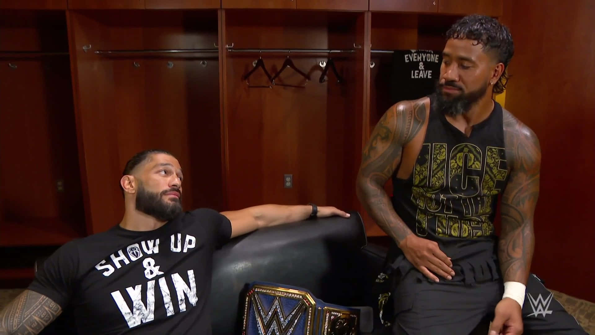 WWE 明星：傑伊·烏索與羅曼·雷恩斯沙發合照 壁紙 — 兩名有紋身的WWE摔角手坐在木製更衣室裡，其中一人靠在冠軍腰帶旁邊的沙發上。