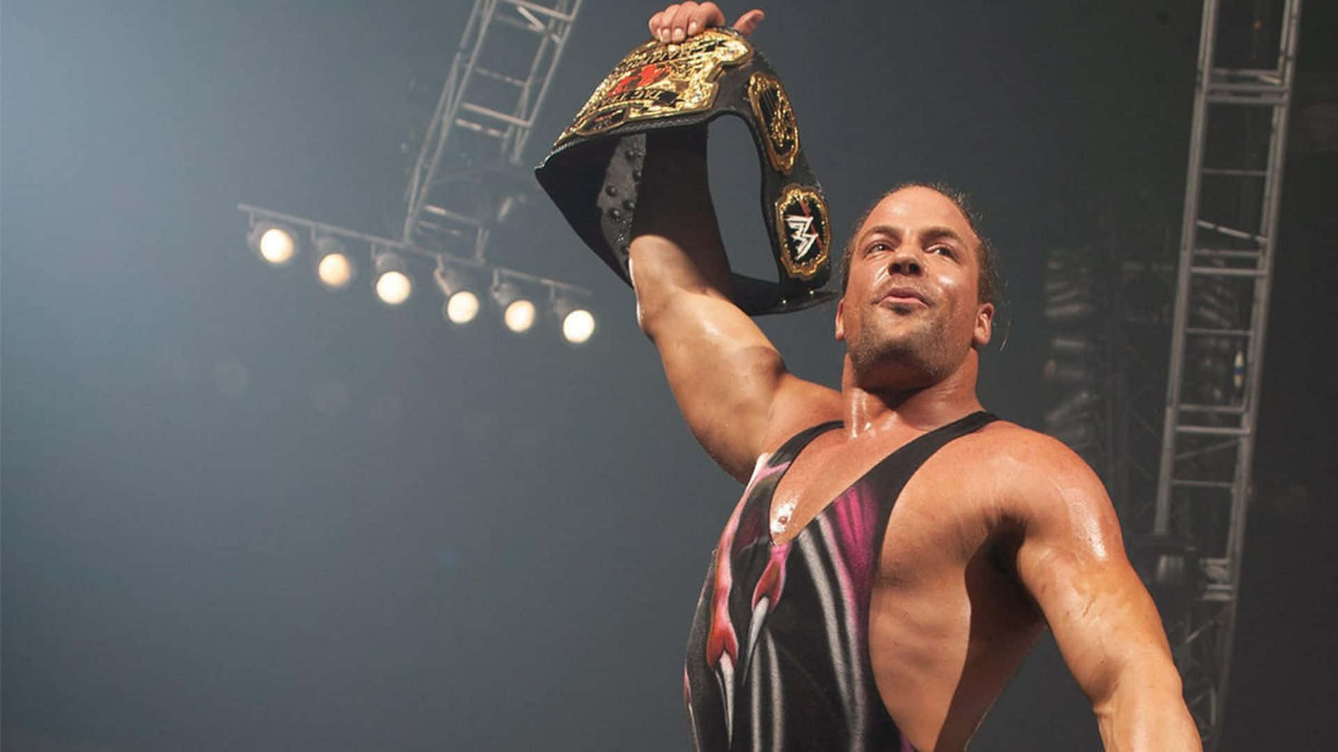 WWE 羅布·范·達姆持冠軍腰帶照 壁紙 — 一位肌肉發達的摔角手在明亮的場館燈光下勝利地高舉冠軍腰帶。