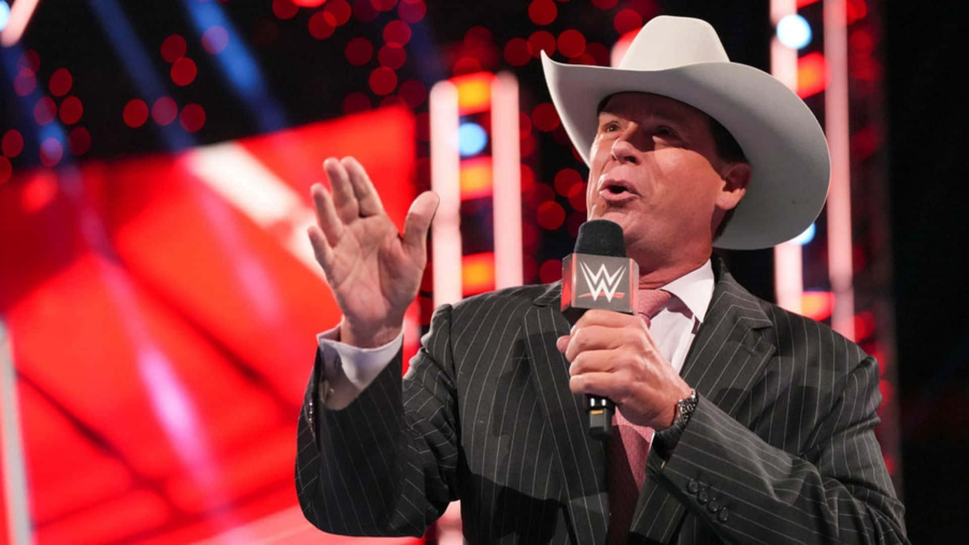 WWE 職業摔角手約翰·布拉德肖·萊夫利賽事動作照 壁紙 — 一個戴著牛仔帽、身穿細條紋西裝的男人，在燈光明亮的舞台上熱情地做手勢，手握WWE麥克風。