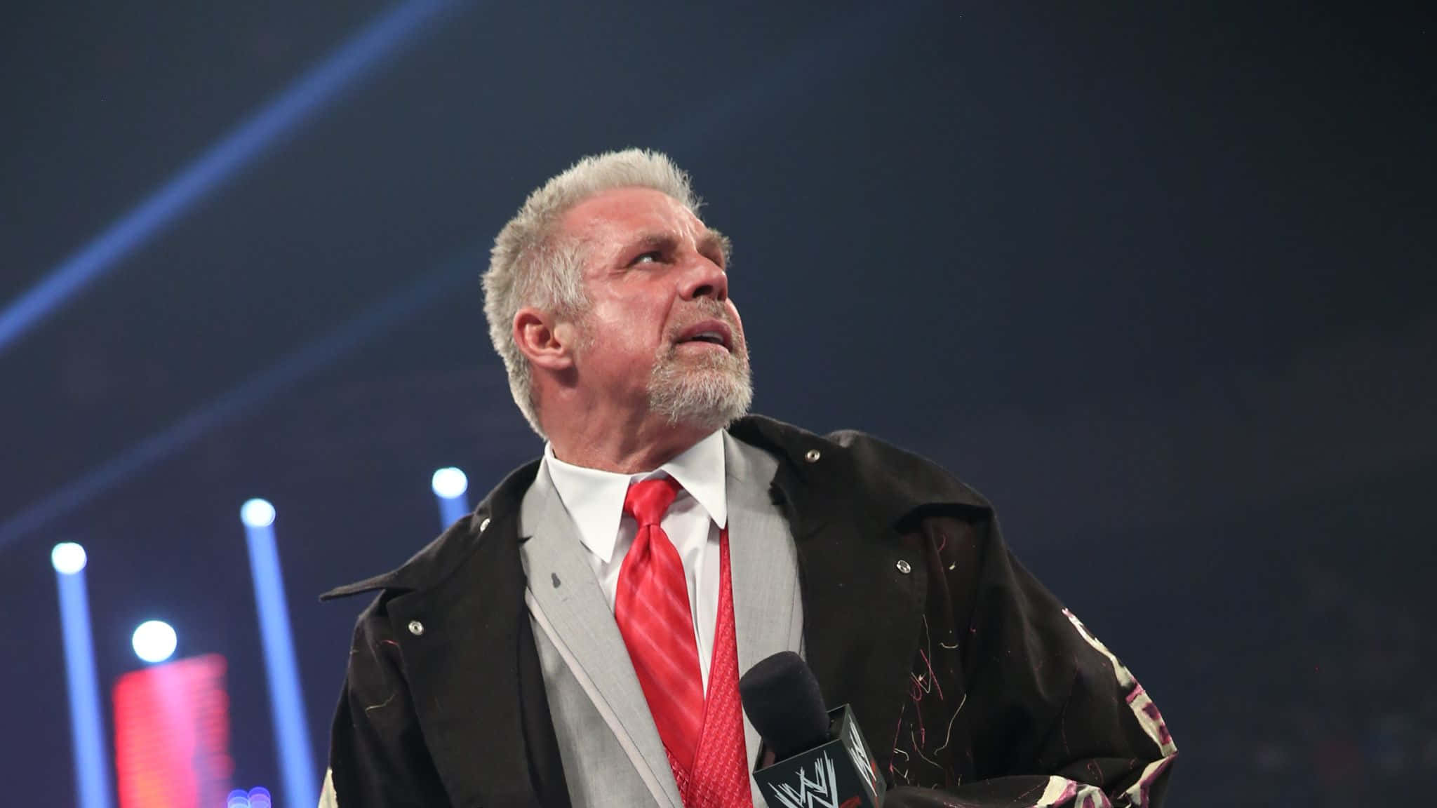 WWE傳奇 Ultimate Warrior 2014 RAW 活動低角度鏡頭 壁紙 — 一位灰白頭髮和鬍鬚的摔角手站在燈光璀璨的舞台上，身穿黑色外套、灰色西裝和紅色領帶，手持WWE麥克風。