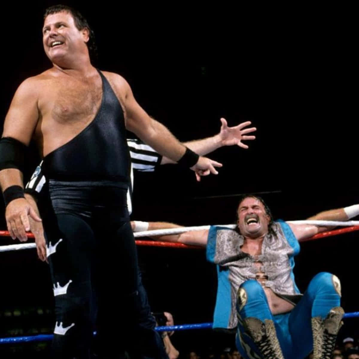 WWE傳奇人物傑克·羅伯茨與傑瑞·勞勒環內對峙 壁紙 — 一名穿著黑色裝備的摔角手，得意地嘲笑著在墊子上痛苦尖叫的藍色裝備對手。