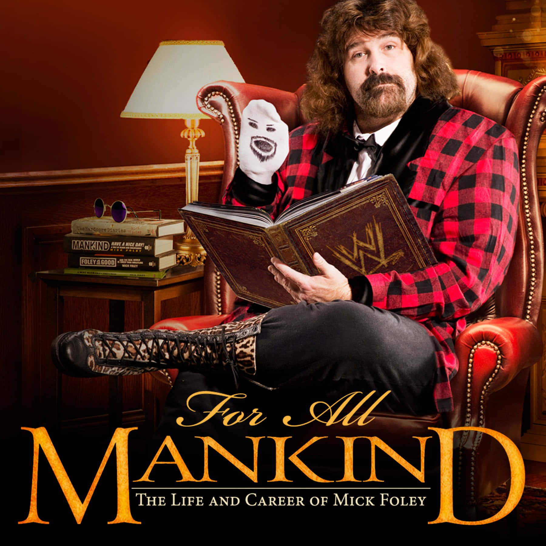 WWE傳奇人物米克·福萊《為所有人類》DVD封面 壁紙 — Mick Foley 坐在紅色皮椅上，手持一本 WWE 書籍和一個白色面具，身穿紅色格紋外套。