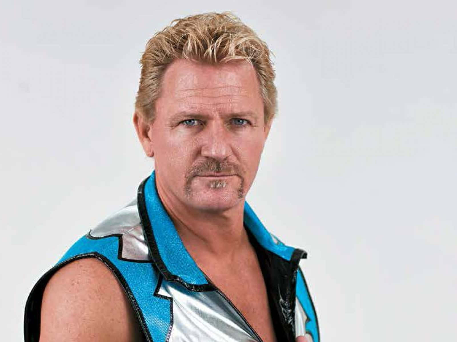 WWE傳奇職業摔角手Jeff Jarrett 壁紙 — 一個肌肉發達、金髮、留鬍子的男人，穿著閃亮的藍銀色摔角背心，背景是純白色。