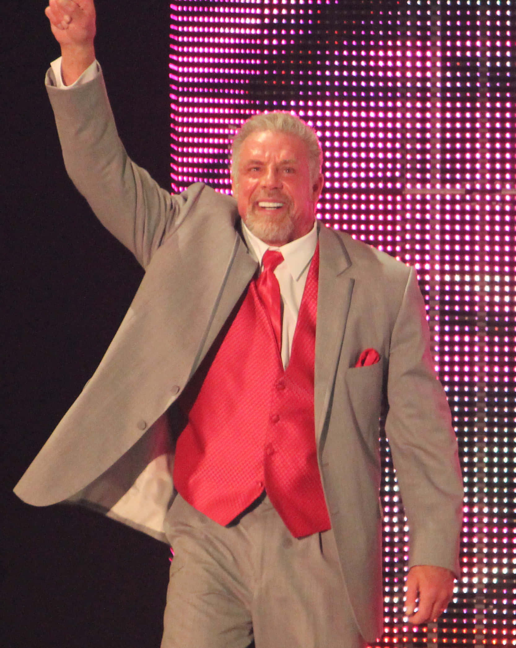 WWE名人堂成員 Ultimate Warrior 2014 照片 壁紙 — 一位穿著灰色西裝和紅色背心的魅力摔角手，在發光的粉紅色LED背景前勝利地舉起拳頭。