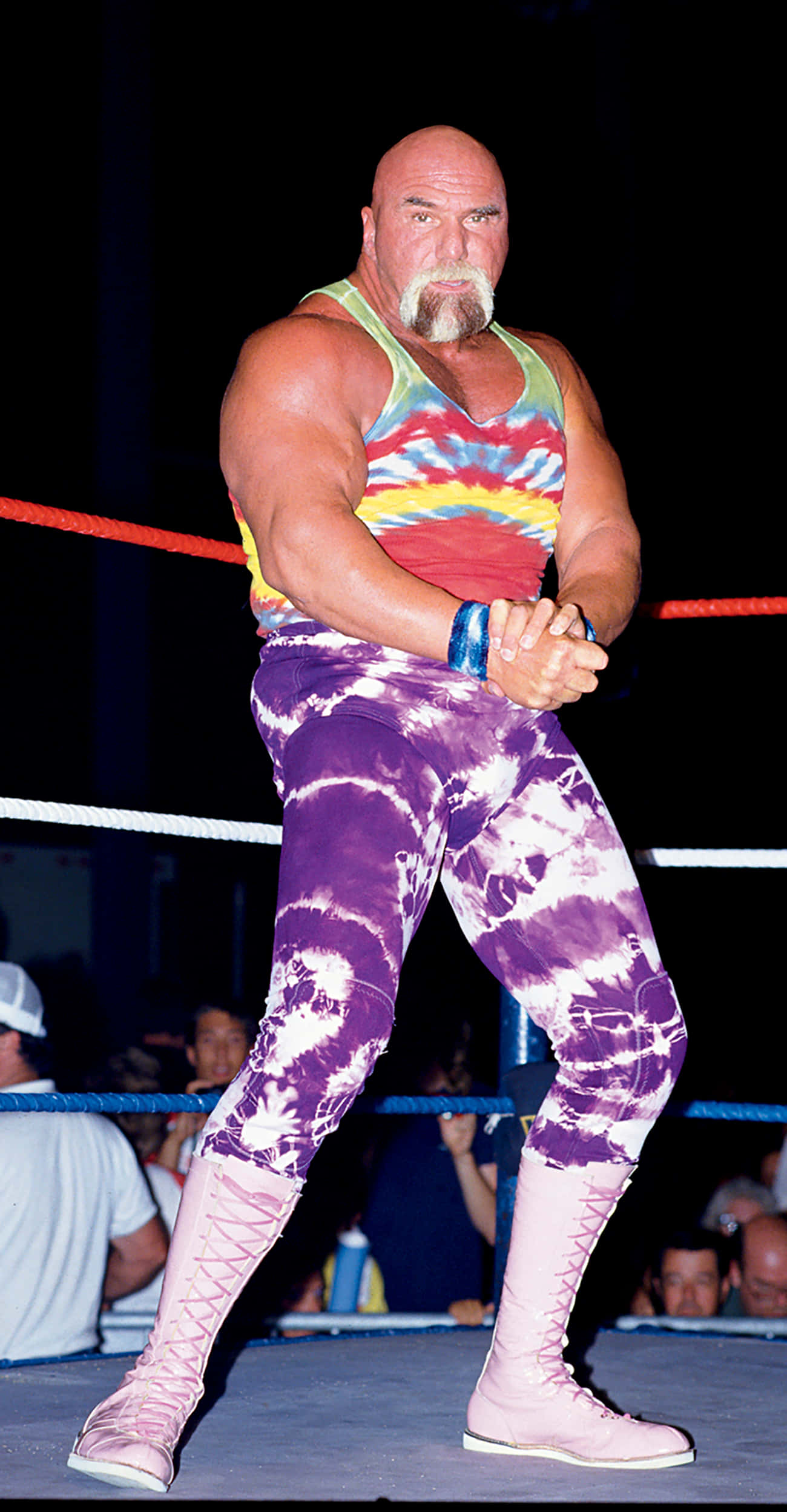 WWE名人堂成員比利·格雷厄姆全身照 壁紙 — 一個肌肉發達的光頭摔角手留著山羊鬍，站在擂台上，穿著色彩繽紛的紮染背心和紫色紮染褲子，配粉紅色靴子。