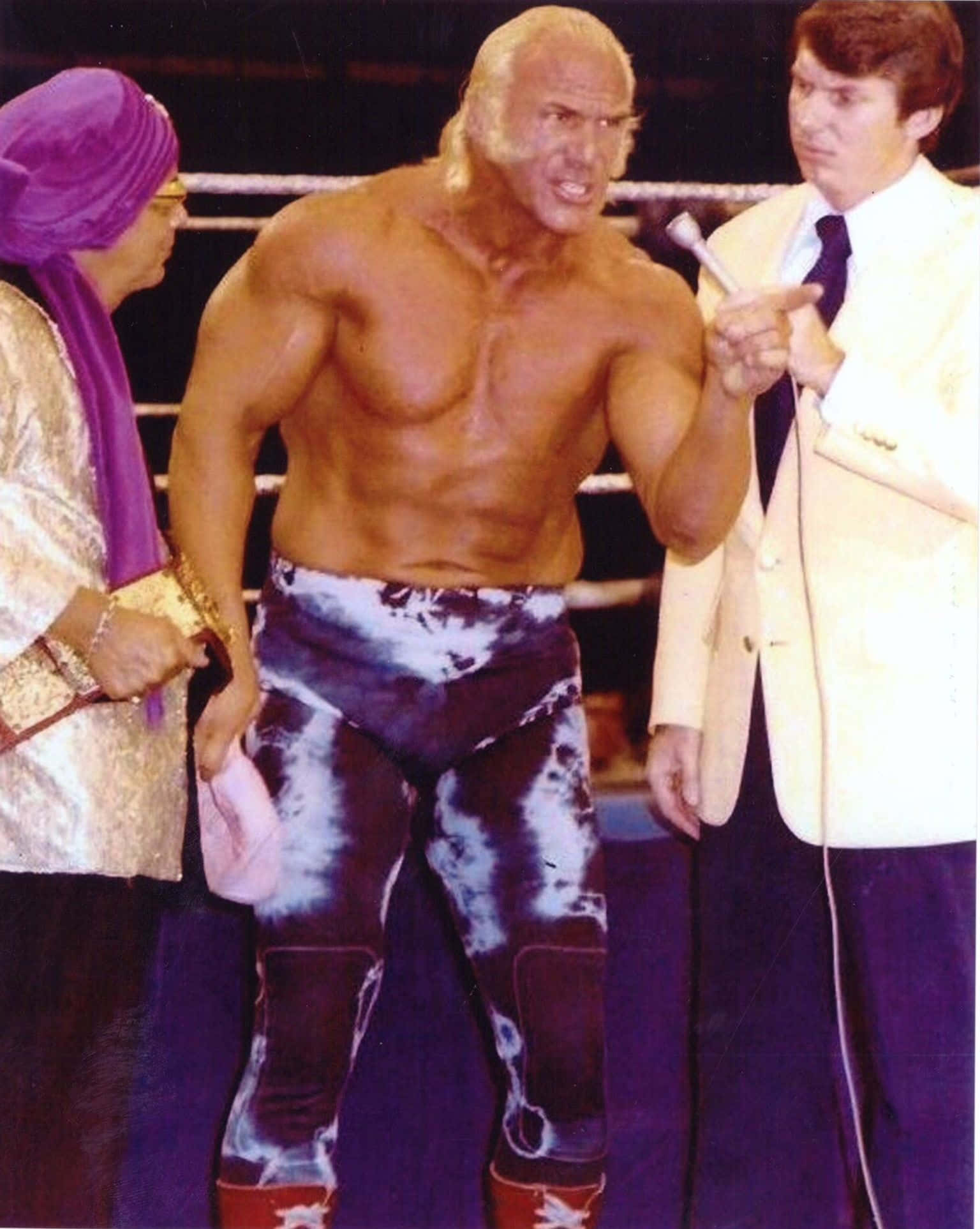 WWE名人堂成員比利·格雷厄姆與文斯·麥馬漢合照 壁紙 — 一位肌肉發達的摔角手穿著紮染短褲站在擂台上，兩旁是一位穿西裝的男人和一位戴紫色頭巾的女人。