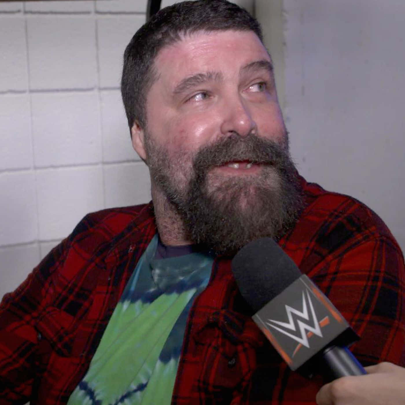 WWE名人堂米克·福萊接受WWE Raw專訪 壁紙 — 一個留鬍鬚的男人，身穿紅色格仔恤衫，對著WWE咪高峰講話，背景是白色瓷磚牆。