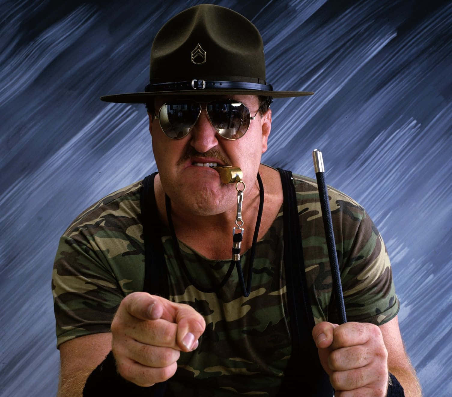 WWE摔角大使Sgt Slaughter 壁紙 — 一位嚴厲的訓練軍士身穿迷彩服和帽子，嘴裡含著哨子，指向前方，背景是帶有條紋的藍色。