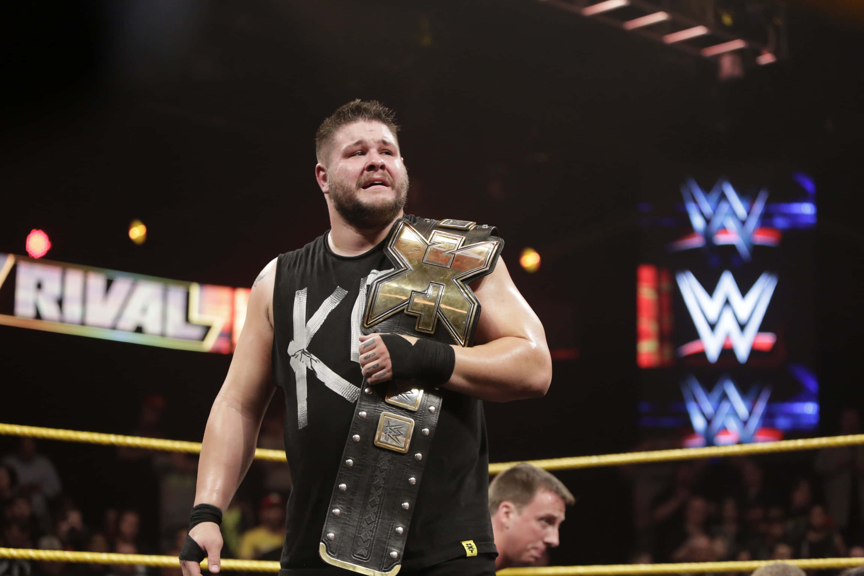 WWE摔角手 Kevin Owens 舉起 WWE NXT 標誌性冠軍腰帶 壁紙 — 摔角手帶著冠軍腰帶，在明亮的場館燈光下於擂台上勝利地站立。