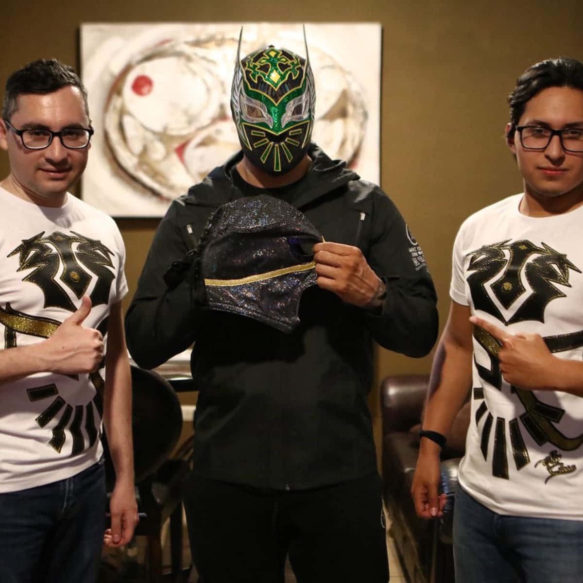 三個室內男子擺姿勢，一人戴著 lucha libre 面具手持另一個面具，兩人穿著相配的龍 T 恤豎起大拇指。