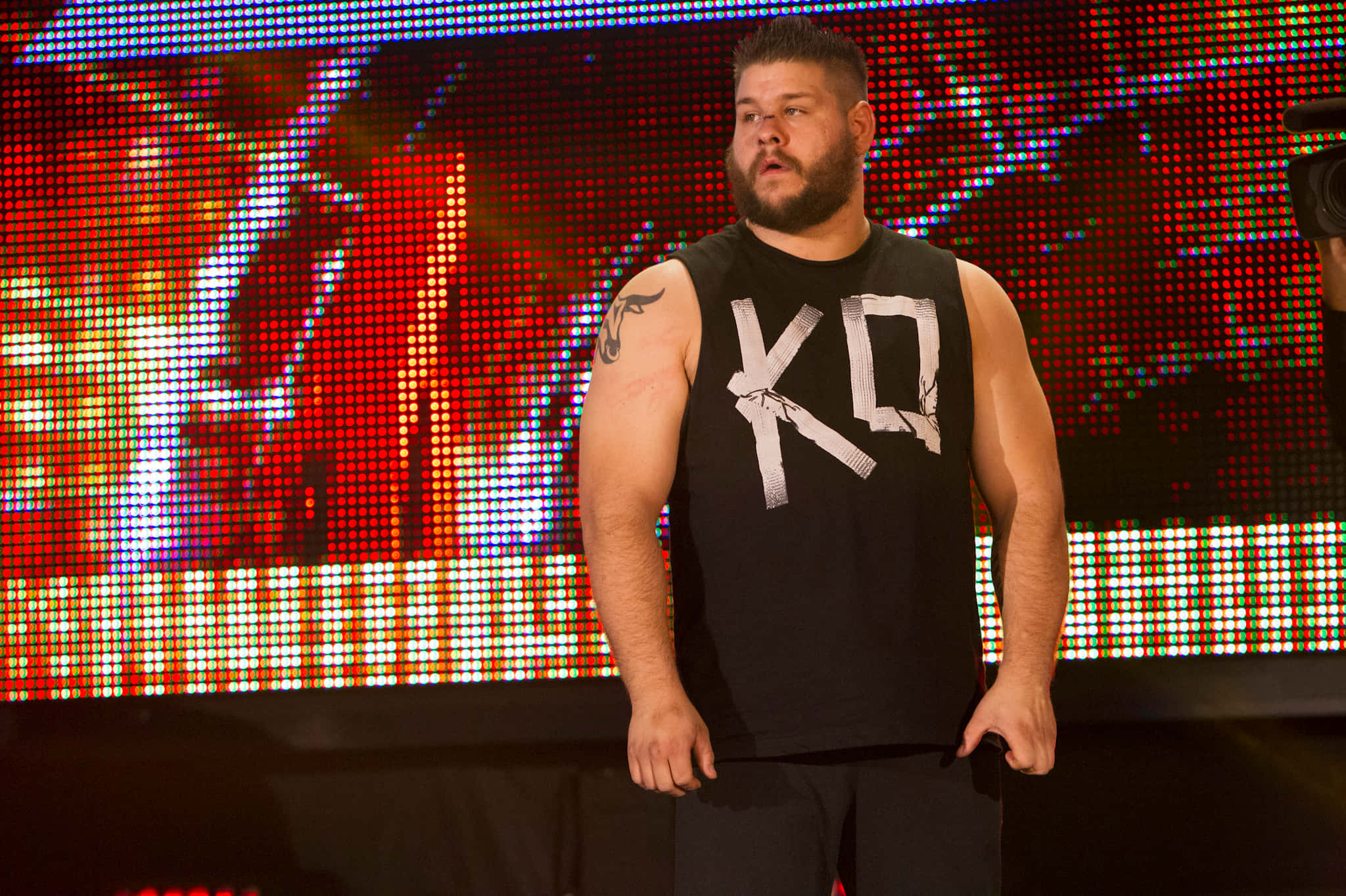 WWE職業重量級摔角手 Kevin Owens 壁紙 — 一位肌肉發達、留鬍子、有紋身的摔角手自信地站在舞台上，面前是充滿活力的LED屏幕。