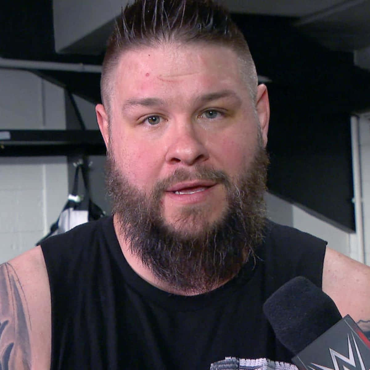 WWE超級巨星 Kevin Owens 的緊湊特寫 壁紙 — 一位留鬍鬚的男人穿著黑色背心，在後台採訪環境中對著WWE麥克風說話。