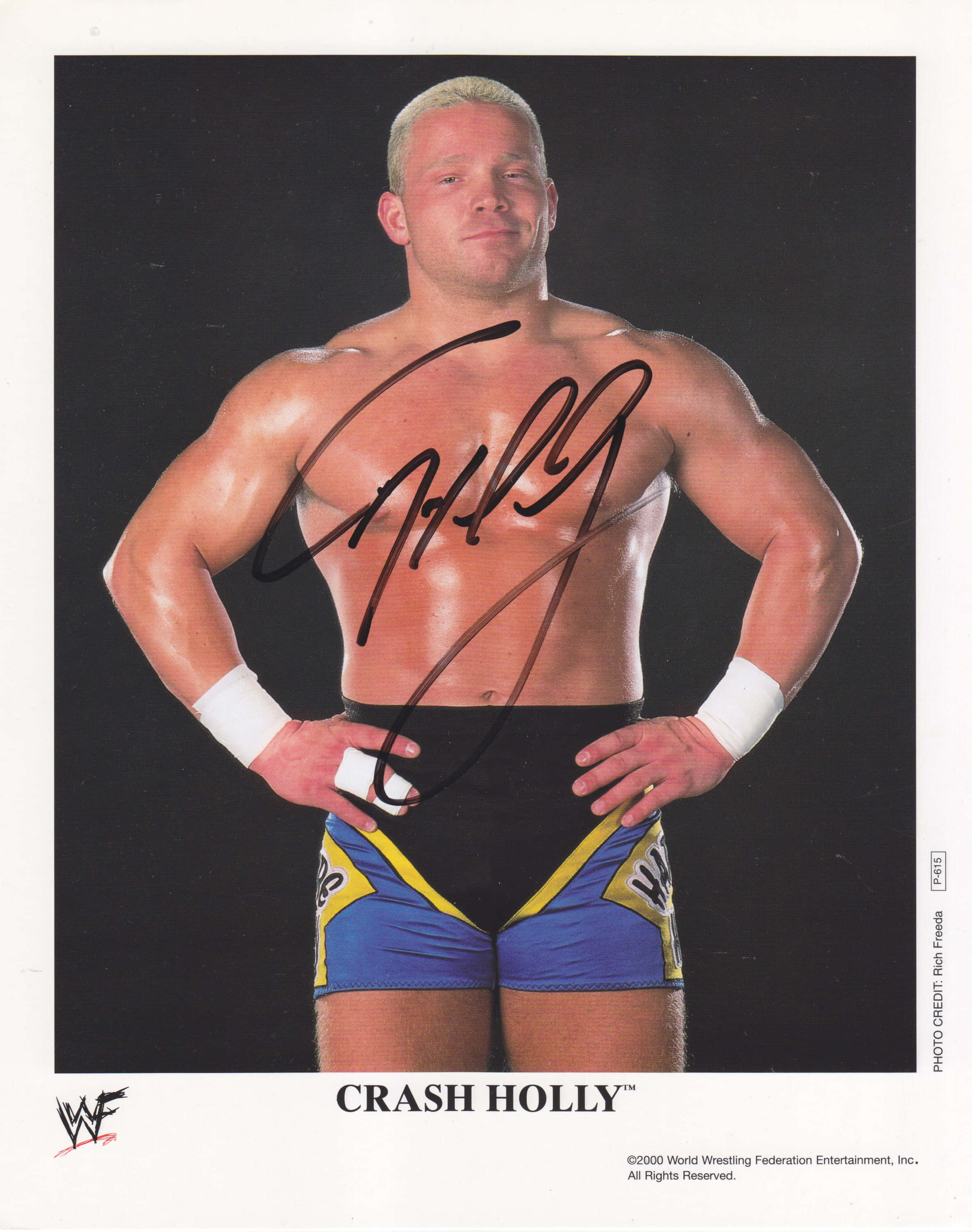 WWE超級巨星克雷什·霍利宣傳照 壁紙 — 肌肉發達的摔角手 Crash Holly 自信地雙手叉腰站在黑色背景前，穿著藍黃色短褲和白色護腕。