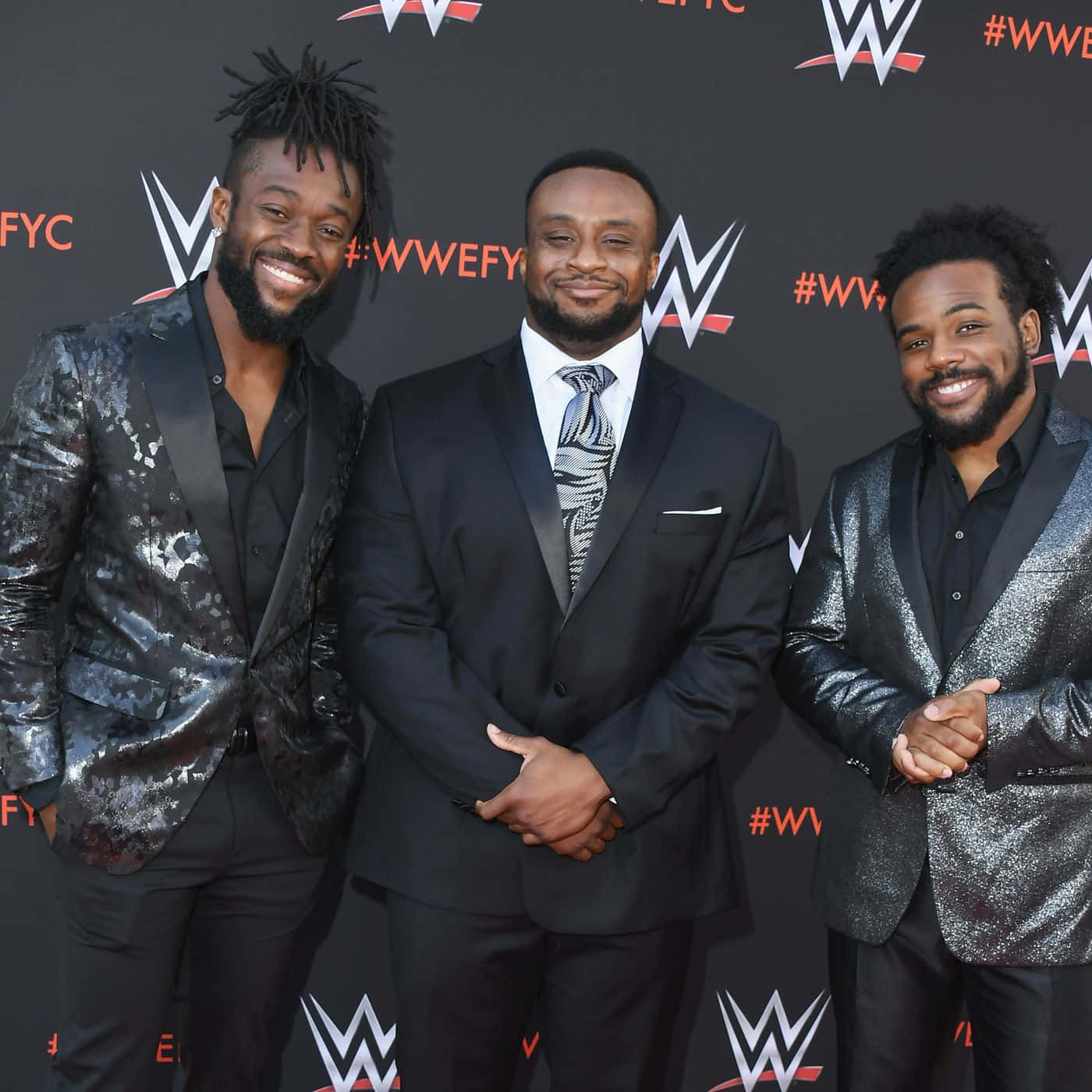 WWE超級巨星科菲·金斯頓與The Big E及Xavier Woods合影 壁紙 — 三位微笑的WWE摔角手穿著華麗西裝，在品牌活動背景前自信擺姿勢。