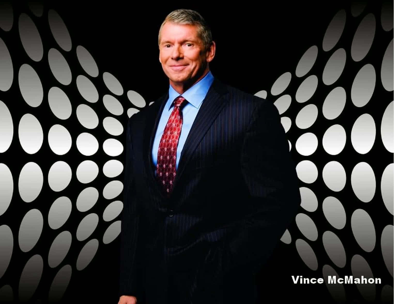 Vince McMahon 身穿深色西裝和紅色領帶，站在帶有白色橢圓燈光的黑色牆壁前。