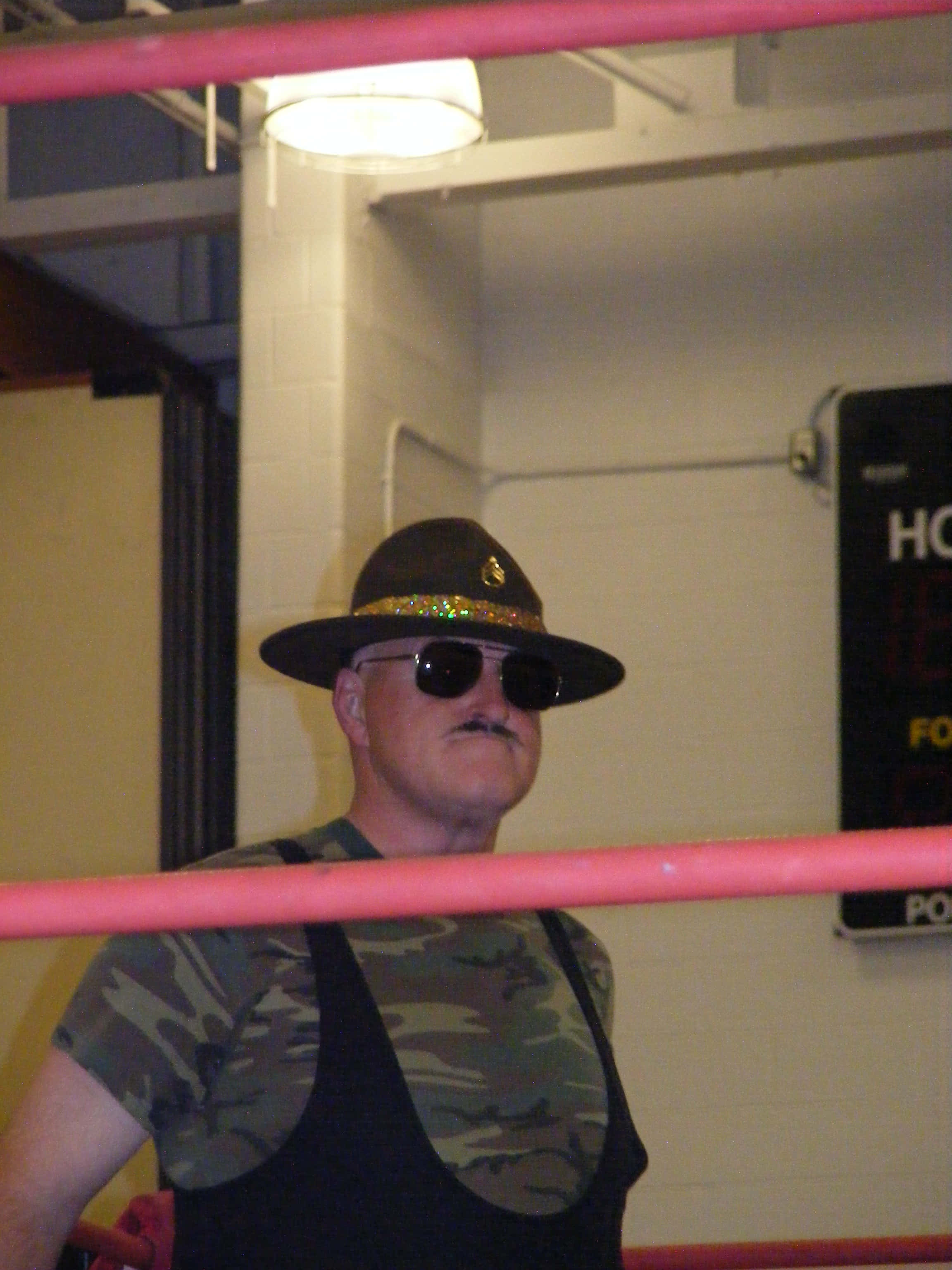 WWF摔角傳奇Sgt Slaughter 壁紙 — 一名身穿迷彩襯衫和寬邊帽的摔角手，在明亮的頭頂燈光下，站在一個粉紅色邊緣的摔角擂台內。