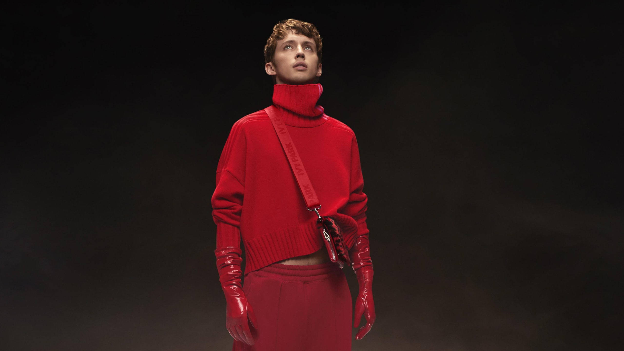 特洛伊·西文 X Ivy Park x 阿迪達斯 壁紙 — 一位模特兒穿著大膽的紅色高領毛衣、手套和裙子，站在黑暗煙霧繚繞的背景前，搭配一個相配的紅色斜孭袋。