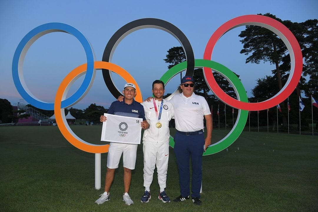 Xander Schauffele 與奧運標誌合影 壁紙 — 三名美國奧運高爾夫球手在黃昏時分，在巨大的奧運五環前自豪地展示金牌和記分卡。