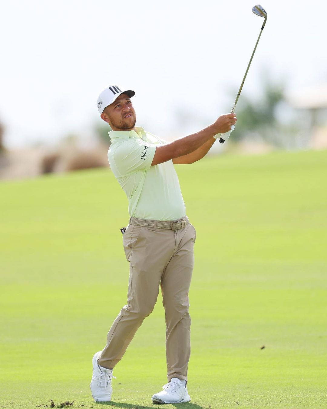 Xander Schauffele 全身揮杆動作 壁紙 — 一名身穿淺綠色襯衫和米色褲子的男性高爾夫球手在晴朗的天空下，在明亮的綠色球道上揮桿。