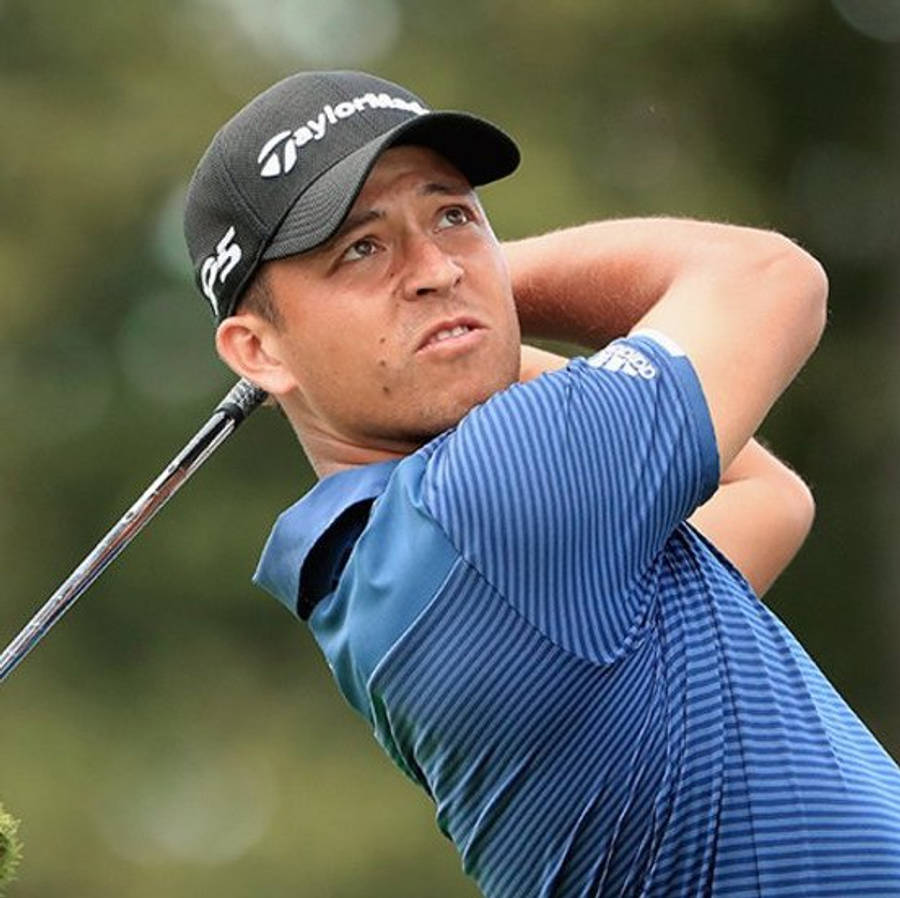 Xander Schauffele 面露嚴肅表情 壁紙 — 一位專注的高爾夫球手正在揮桿，身穿藍色條紋襯衫和黑色帽子，背景是模糊的綠色。