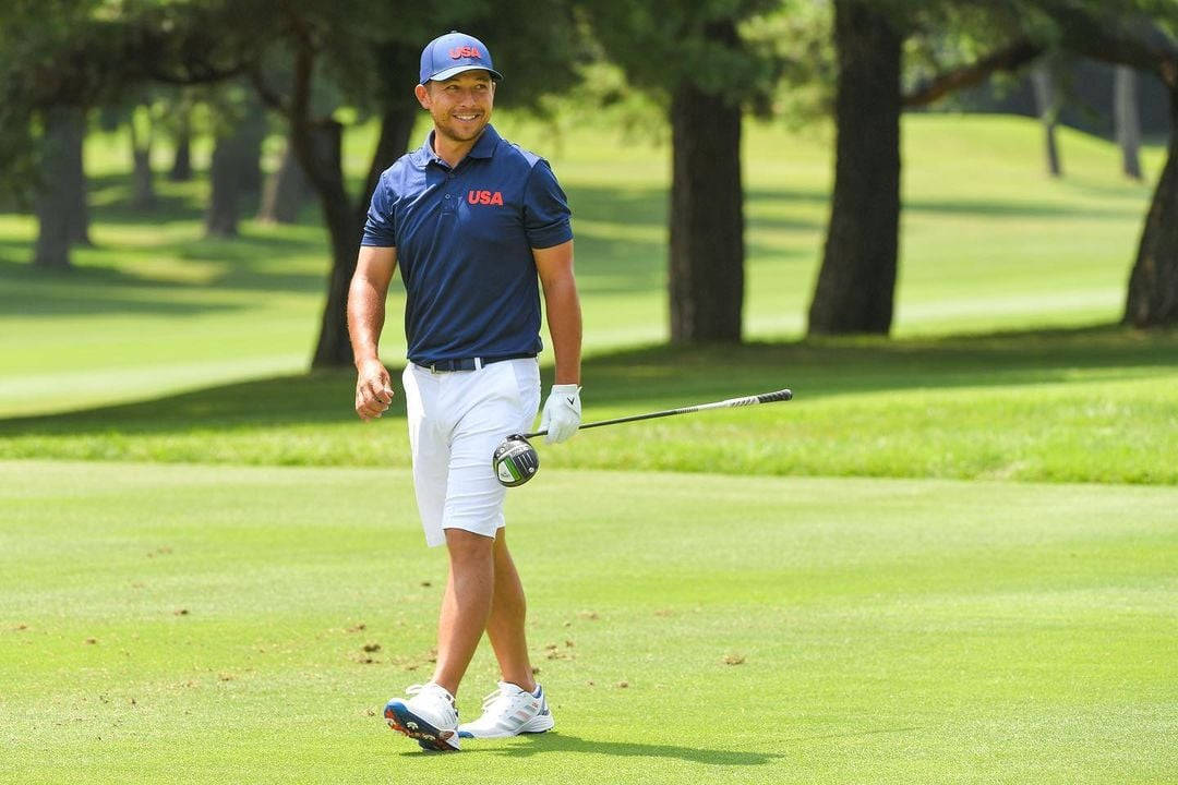Xander Schauffele 持球桿漫步高爾夫球場 壁紙 — 一位微笑的高爾夫球手身穿藍色美國Polo衫和白色短褲，手持球桿走過陽光明媚的綠色球道。