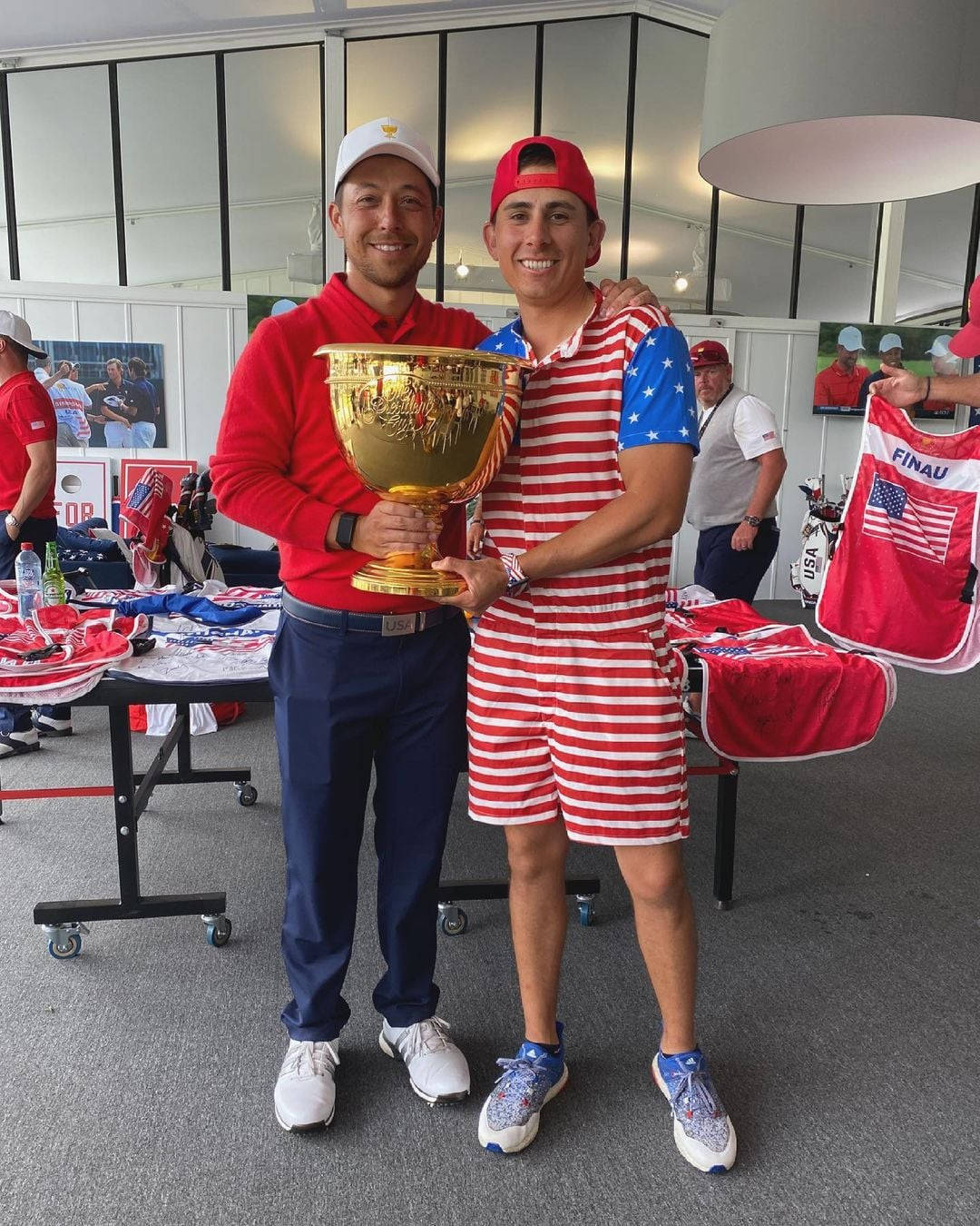 Xander Schauffele 舉起獎盃慶祝勝利 壁紙 — 兩名面帶微笑的高爾夫球手身穿紅、白、藍色的美國裝備，在活動中自豪地共同舉起一個大型金色獎盃。