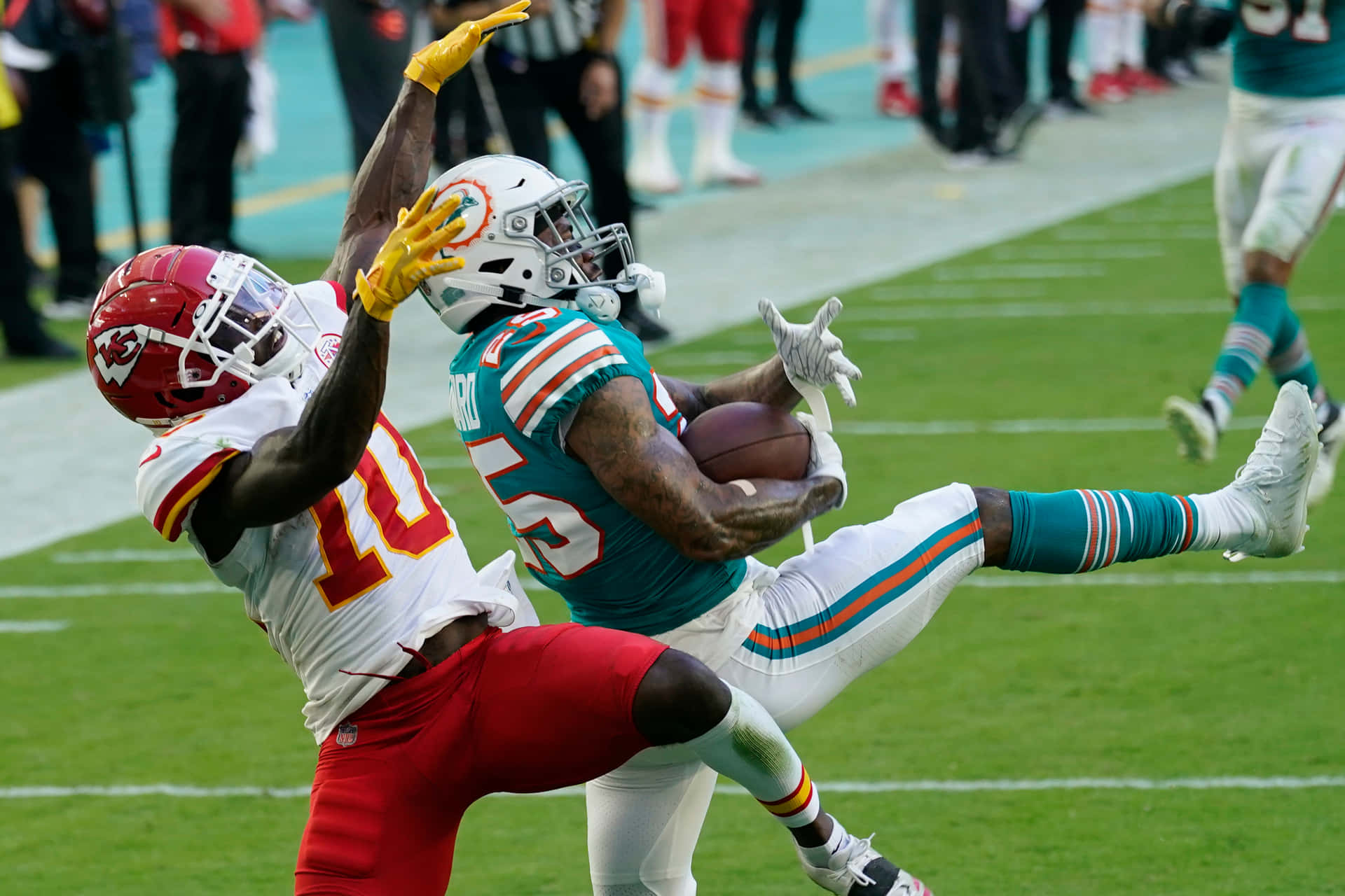 Xavien Howard激烈足球賽 壁紙 — 邁阿密海豚隊的外接手在空中跳起抓住橄欖球，而堪薩斯城酋長隊的防守球員則在充滿活力的綠色球場上飛身擒抱他。