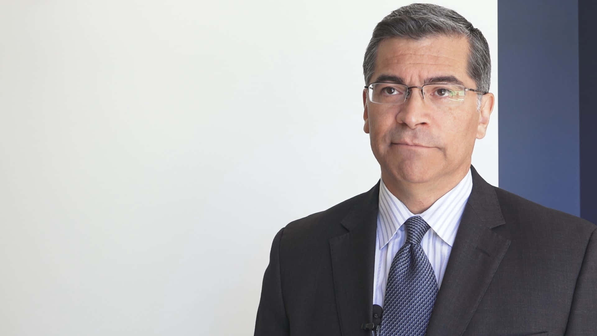 Xavier Becerra 冷靜無表情 壁紙 — 一位戴眼鏡的中年男子穿著深色西裝和圖案領帶，背景是純白色和藍色。