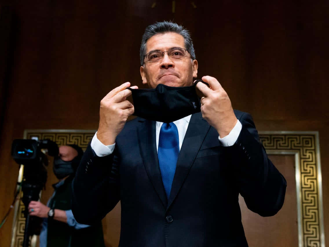Xavier Becerra 摘下口罩 壁紙 — 一名身穿深色西裝和藍色領帶的男子在一個帶有華麗金色飾邊的正式木質房間內取下黑色口罩。