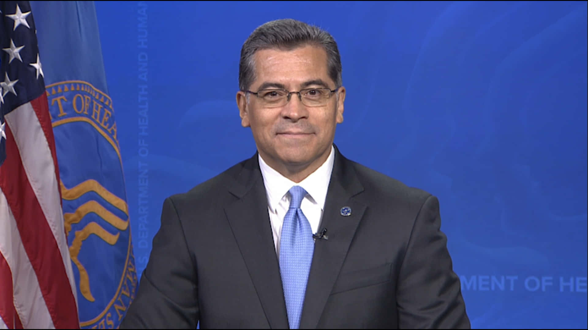 Xavier Becerra 靜謐微笑 壁紙 — 一名身穿西裝和眼鏡的男子站在藍色背景前，背景有美國國旗和衛生與公眾服務部的旗幟。