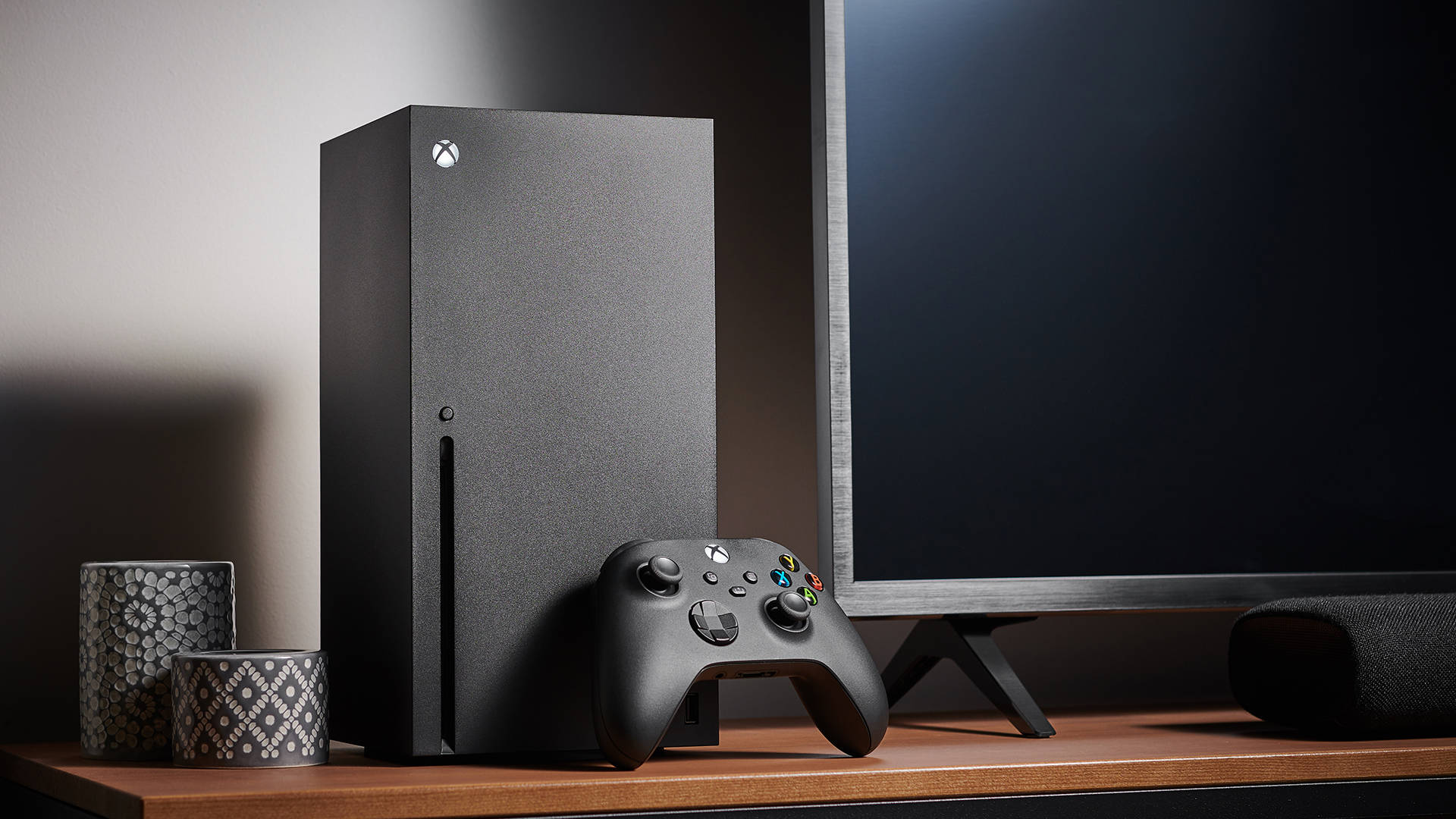 Xbox Series X 控制器擺放桌上 壁紙 — 在柔和的工作室燈光下，一個時尚的黑色 Xbox Series X 主機和控制器放在木架上，旁邊有電視和圖案燭台。