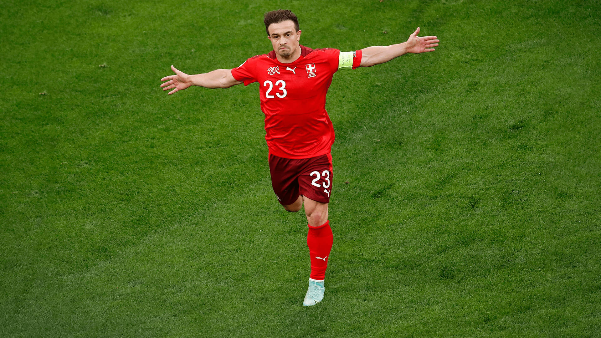 Xherdan Shaqiri 足球勝利慶祝 壁紙 — 一位身穿紅色23號球衣的瑞士足球運動員在充滿活力的綠色球場上張開雙臂慶祝。