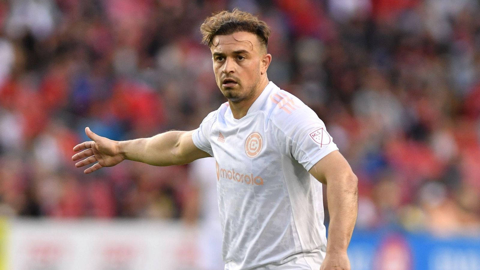 Xherdan Shaqiri 世界盃賽場瞬間 壁紙 — 一位專注的足球員身穿白色球衣，在比賽中做出手勢，背景是模糊的人群。