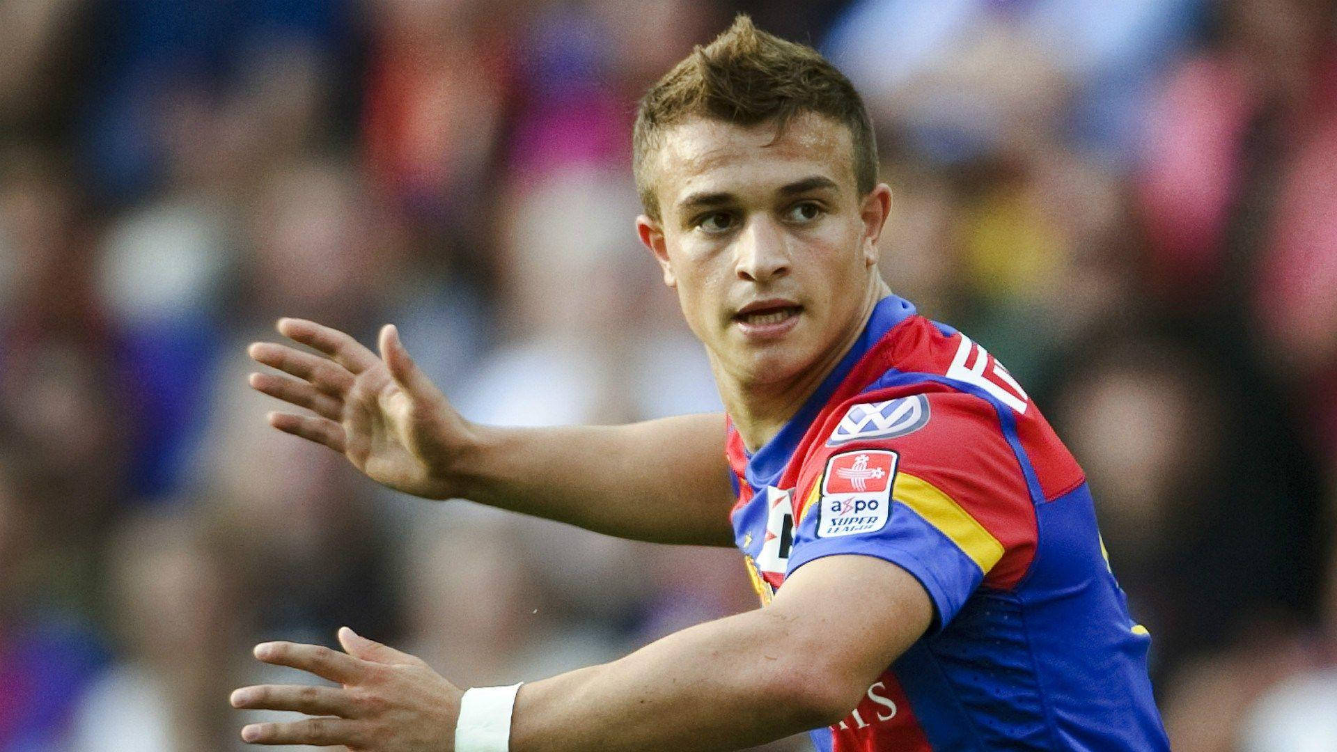 Xherdan Shaqiri 瑞士足球員專注神情 壁紙 — 一位專注的足球員身穿紅藍球衣，在日間比賽中動態地做手勢，背景是模糊的觀眾。