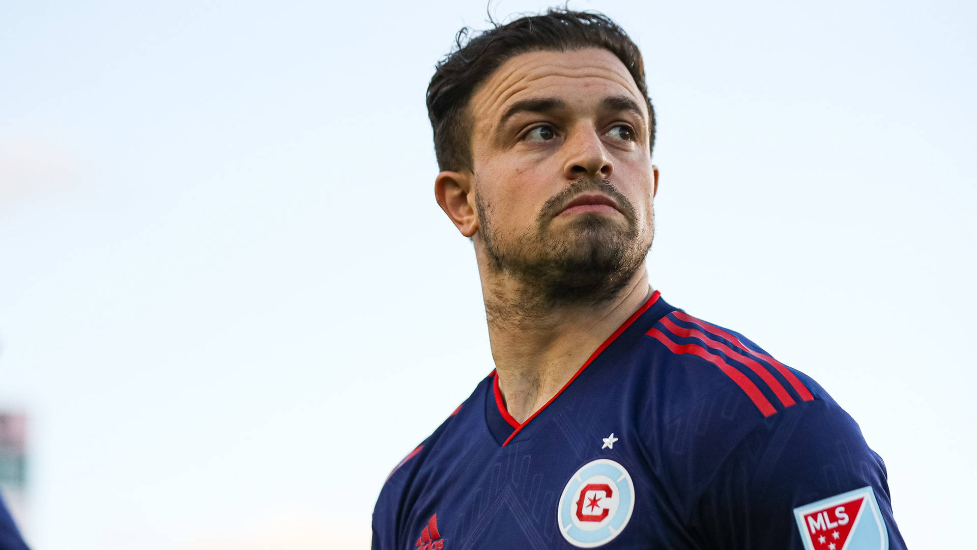 Xherdan Shaqiri 職業足球員靜態肖像 壁紙 — 一位專注的足球運動員身穿海軍藍色帶紅色條紋的球衣，仰望明亮的天空。