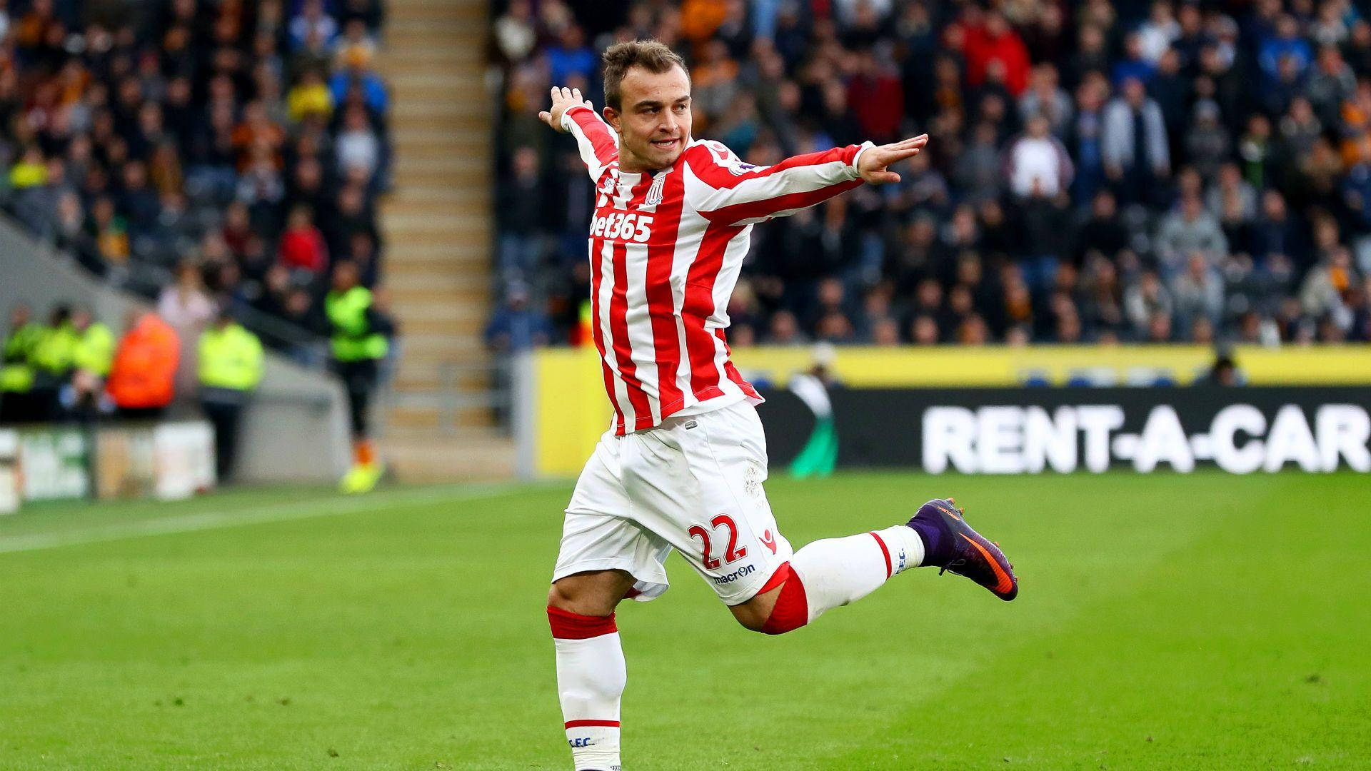 Xherdan Shaqiri 足球賽場搞笑瞬間 壁紙 — 一名身穿紅白間條球衣的足球員在綠色球場上張開雙臂慶祝入球，背景是模糊的人群。